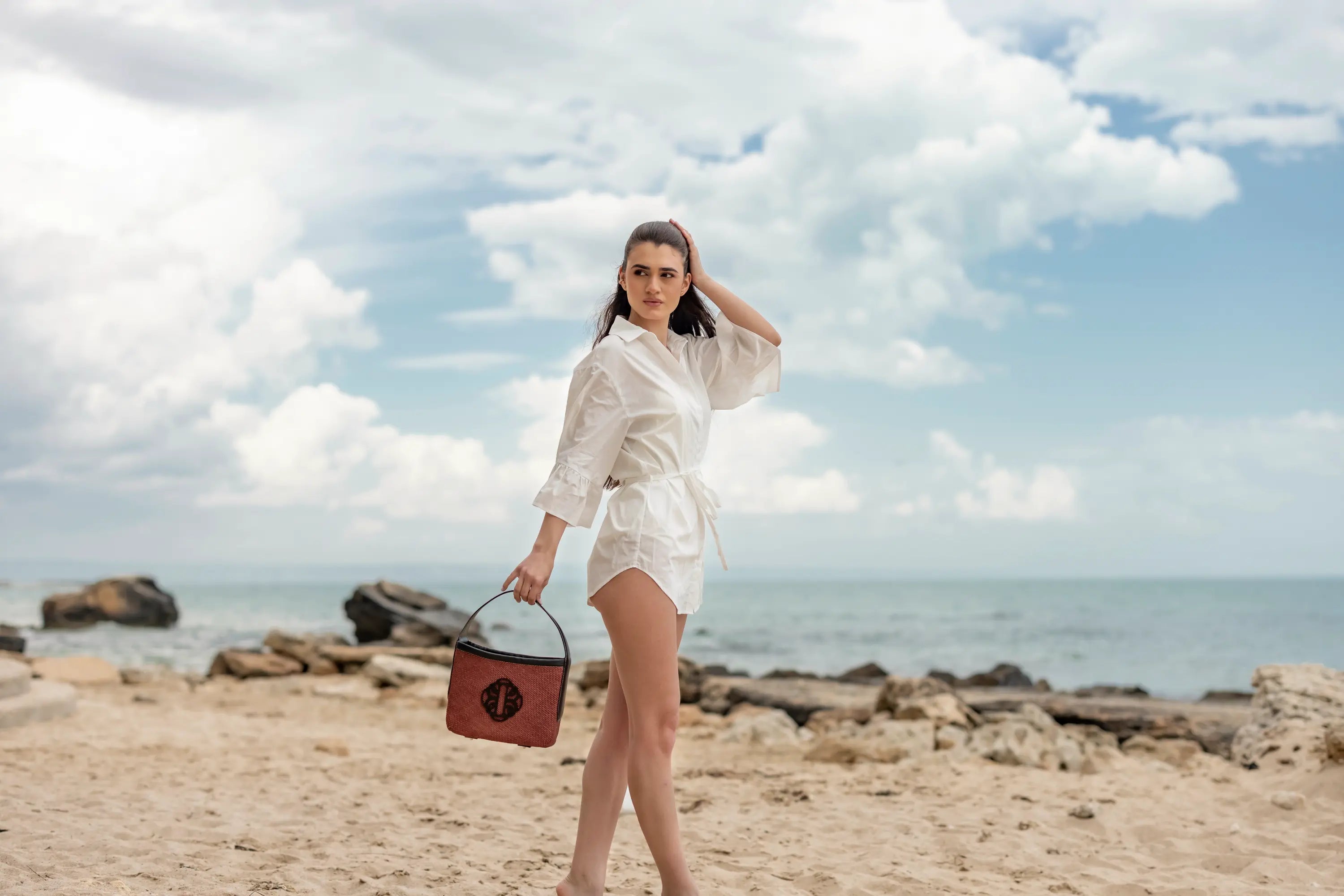 Woman in a white outfit holding a red bag on a beach with ocean and sky in the background