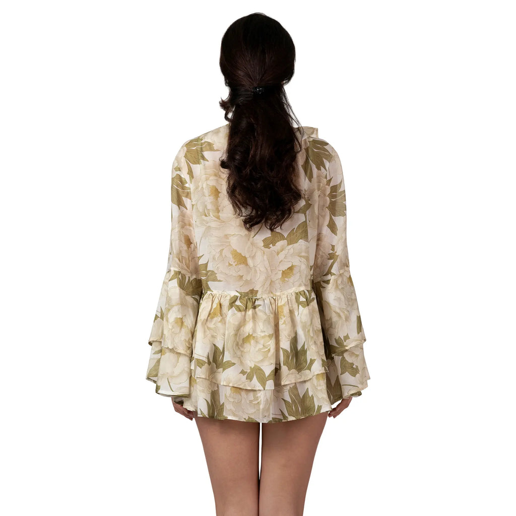 Woman wearing the FLEUR DE LUMIÈRE Short Tunic by BILEYA COUTURE, crafted from luxurious silk-cotton chiffon with a romantic white peony floral print, voluminous ruffle sleeves, and an airy designer resort wear silhouette.
